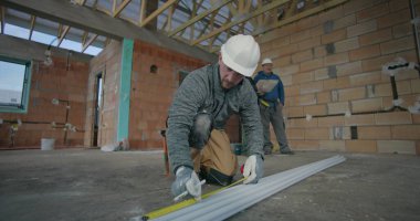 Meslektaşlar planlarını gözden geçirirken, inşaat işçisi borular üzerinde mezura kullanırken, bina inşaatında takım çalışması ve hassasiyet içeren kapalı inşaat ortamı da kullanılıyor.