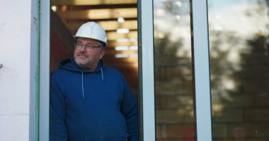 Kapıda duran inşaat işçisi, kolları rahat, başlık takıyor, inşaat çalışmalarını dikkatle izliyor, inşaat alanında deneyimli bir müteahhit iş başında.