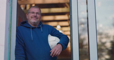 Emniyet kaskı takmış, güvenle gülümseyen, kapı aralığına yaslanan, inşaat sektöründe profesyonel inşaat projesiyle uğraşan bir işçi.