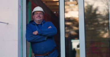 Kapıda duran inşaat işçisi, kollar çapraz, güvenlik teçhizatı giyiyor, inşaat sürecini denetliyor, kendine güvenen duruş, inşaat yönetimi ve denetim