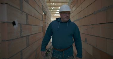 İnşaat alanının koridorunda yürüyen, güvenlik kaskı ve alet kemeri takan, tuğla duvarları ve ahşap tavanı olan bir erkek işçi.