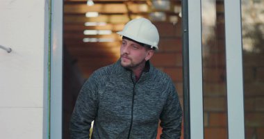 Kapıda duran inşaat işçisi, düşünceli bir şekilde mesafeye bakıyor, kafasında şapka, tuğla duvar arka planı, endüstriyi, işçiliği ve işçiliği temsil ediyor.