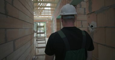 Erkek işçi, tamamlanmamış binanın koridorunda yürüyor, miğfer takıyor, etrafı inceliyor, tuğla duvarlar ve ahşap iskeletle kaplı.