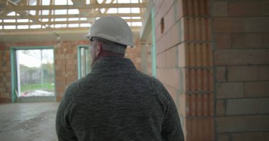 İnşaat işçisi miğferiyle bina içini teftiş ediyor, dikiz aynasından bakıyor, ahşap merdiveni ve tamamlanmamış tuğla duvarları aşıyor.