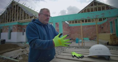 İnşaat alanında eldiven ve miğfer takan bir adam, arka planda kısmen inşa edilmiş bir evin yanında duruyor, dışarıda çalışmaya hazırlanıyor.