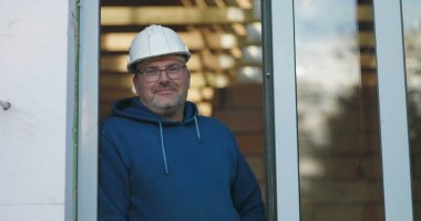 Kapıda duran bir işçi, hafif bir gülümseme, güvenlik kaskı ve gözlük takan, gururlu ama profesyonel bir tavırla inşaat projesini denetleyen