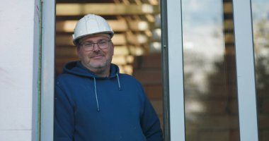 Kapıda duran bir işçi, hafif bir gülümseme, güvenlik kaskı ve gözlük takan, gururlu ama profesyonel bir tavırla inşaat projesini denetleyen