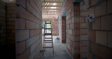 Açığa çıkmış tuğla duvarları, ahşap tavan kirişleri ve kapı eşiğine yerleştirilmiş bir merdiveni olan tamamlanmamış binanın iç manzarası.