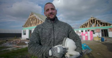 İnşaat alanının önünde kaskını tutan adam, inşaat halindeki evlerle güvenle ayakta duruyor, eldiven takıyor, doğrudan kameraya bakıyor.