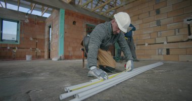 Meslektaşlar planlarını gözden geçirirken, inşaat işçisi borular üzerinde mezura kullanırken, bina inşaatında takım çalışması ve hassasiyet içeren kapalı inşaat ortamı da kullanılıyor.