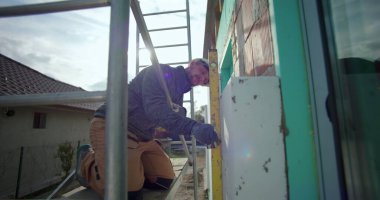 Dış duvardaki izolasyon panelini hizalamak için ruh seviyesini kullanmak. İnşaat işçisi, inşaat iskelesine dizlerinin üzerine çöküp, güvenlik teçhizatı, eldivenler, konut binası.