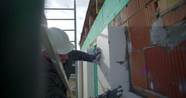 Tuğla duvara yalıtım paneli dayanıyor, inşaat işçisi arka planda güvenlik eldivenleri, miğfer ve merdiven ile yalıtım düzenliyor.