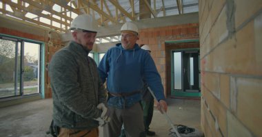 İnşaat işçileri inşaat planlarını tartışıyor. Üç işçi, inşaat başlıkları ve güvenlik eldivenleri takıyor. Bitmemiş bina planlarını gözden geçiriyorlar.