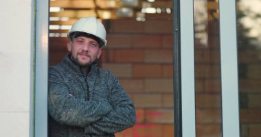 Kollarını kavuşturmuş inşaat işçisi, güvenli bir şekilde mesafeye bakıyor, başlık takıyor, kapının önünde duruyor, gücü, güvenilirliği ve kalifiyeli işçiliği simgeliyor.