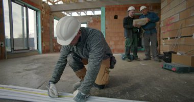 İnşaat işçisi borularda şerit kullanırken, iki işçi arka planda planları, inşaat halindeki kapalı inşaat alanını, takım çalışmasını ve hassasiyetini tartışıyor.