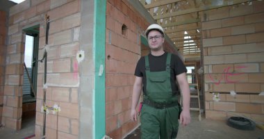 İnşaat alanında yürüyen, miğfer ve yeşil tulumlar giyen, tamamlanmamış tuğla duvarları ahşap tavan iskeletiyle inceleyen bir erkek işçi.