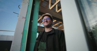 Sert şapka ve gözlüklerle gülümseyen inşaat işçisi, inşaat alanında açık çatı çerçevesi altında güvenle duruyor, pozitif tavır sergiliyor ve projeye odaklanıyor