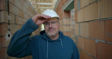 Erkek işçi bitmemiş binanın koridorunda duruyor, güvenlik kaskı ve gözlük takıyor, tuğla duvarları açık, doğrudan kameraya bakıyor.