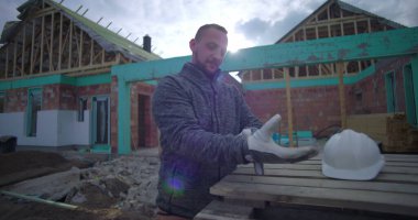 İnşaat alanında eldiven ve miğfer takan adam, arka planda kısmen inşa edilmiş evle ahşap paletlerin yanında duruyor, dışarıda çalışmaya hazırlanıyor.