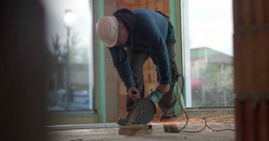 İnşaat işçisi çimento üzerinde açı öğütücü kullanıyor, kıvılcımlar yaratıyor, malzemeyi kesiyor, koruyucu kask takıyor, yoğun konsantrasyon, iç inşaat işi yapıyor.