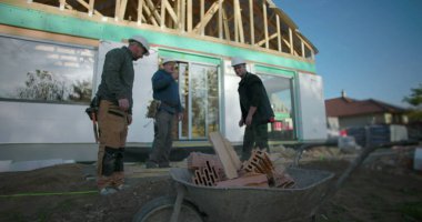 Sert şapkalı inşaat işçileri el arabasına tuğla fırlatıyor, kısmen inşa edilmiş ahşap iskeletli bir evin dışında inşaat projesinde işbirliği yapıyorlar.