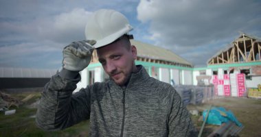 Başlık takmak, kameraya gülümseyen inşaat işçisi, arka planda inşaat alanı, profesyonel ve cana yakın görünüm.