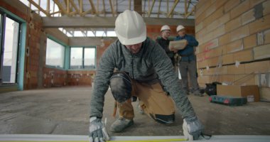 İnşaat işçisi borularda şerit kullanırken, iki işçi arka planda planları, inşaat halindeki kapalı inşaat alanını, takım çalışmasını ve hassasiyetini tartışıyor.