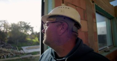 Sert şapkalı, açık havada duran, kapının yanında duran, arka planda doğal çevresi ve enkazı olan olgun inşaat işçilerinin yan profili.