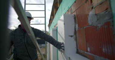 Tuğla duvara yalıtım paneli dayanıyor, inşaat işçisi arka planda güvenlik eldivenleri, miğfer ve merdiven ile yalıtım düzenliyor.