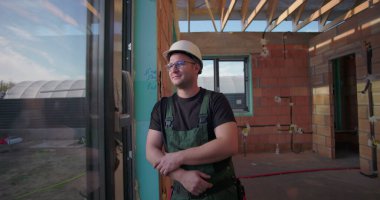 Kapıya dayanmış genç bir inşaat işçisi, sert bir şapka ve yeşil tulum giymiş, bitmemiş bir binada sakin bir ifadeyle dışarıya bakıyor.