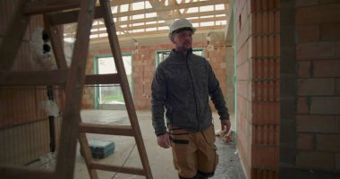 İnşaat işçisi miğferiyle inşaat halindeki evde yürüyor, ilerlemeyi gözlemliyor, etrafı tuğla duvarlar, ahşap kirişler ve pencere çerçeveleriyle çevrili.