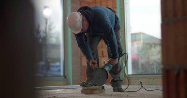 İnşaat işçisi çimento üzerinde açı öğütücü kullanıyor, kıvılcımlar yaratıyor, malzemeyi kesiyor, koruyucu kask takıyor, yoğun konsantrasyon, iç inşaat işi yapıyor.