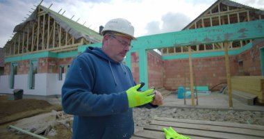 İnşaat alanında eldiven ve miğfer çıkaran, gözlük takan, arka planda inşaat halinde olan bir adam dışarıda çalışmayı bitirmeye hazırlanıyor.