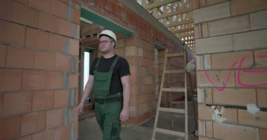 İnşaat alanında yürüyen, miğfer ve yeşil tulumlar giyen, tamamlanmamış tuğla duvarları ahşap tavan iskeletiyle inceleyen bir erkek işçi.