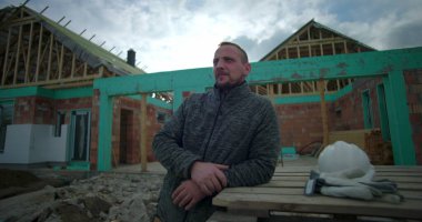 İnşaat alanındaki ahşap paletlere yaslanan adam, yanına yerleştirilmiş kask ve eldivenler, güneş ışığıyla uzaklığa bakıyor, arka planda kısmen inşa edilmiş bir ev.