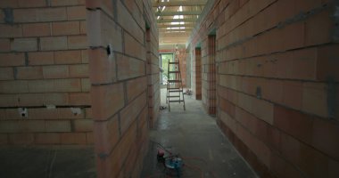 İnşaat halindeki tamamlanmamış evin iç manzarası, açıkta duran tuğla duvarlar, ahşap iskelet ve koridorda bir merdiven.