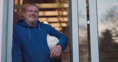 Emniyet kaskı takmış, güvenle gülümseyen, kapı aralığına yaslanan, inşaat sektöründe profesyonel inşaat projesiyle uğraşan bir işçi.