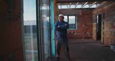 Olgun inşaat işçisi, güvenle gülümsüyor, kasketi takılı, tamamlanmamış tuğla duvarlar ve açık tavan kirişleri ile binanın içinde duruyor.