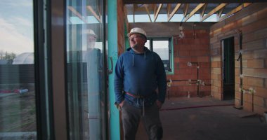 Olgun inşaat işçisi, güvenle gülümsüyor, kasketi takılı, tamamlanmamış tuğla duvarlar ve açık tavan kirişleri ile binanın içinde duruyor.