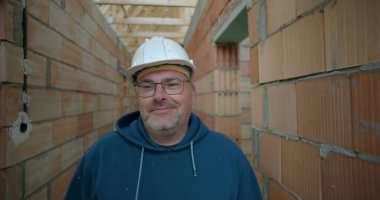 Erkek işçi bitmemiş binanın koridorunda duruyor, güvenlik kaskı ve gözlük takıyor, tuğla duvarları açık, doğrudan kameraya bakıyor.