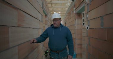 İnşaat alanının koridorunda yürüyen, güvenlik kaskı ve alet kemeri takan, tuğla duvarları ve ahşap tavanı olan bir erkek işçi.