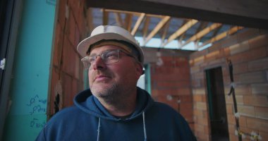 Olgun bir inşaat işçisi, düşünceli bir şekilde bakıyor, kısmen inşa edilmiş binanın içinde, tavan kirişleri ve tuğla duvarları açık.