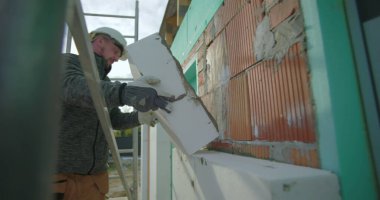 Dış duvara yalıtım paneli yerleştiriliyor, eldiven ve kask takan inşaat işçisi tuğla cephesine baskı yapıyor, konut yenilemesi yapıyor.