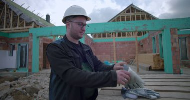 İnşaat alanında eldiven takan, güvenlik kaskı ve gözlük takan, arka planda kısmen inşa edilmiş evi olan ahşap paletlerin yanında duran bir adam.