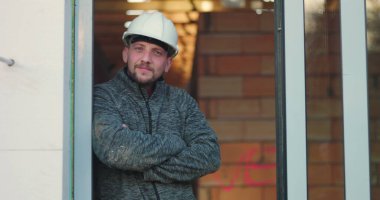 Kollarını kavuşturmuş inşaat işçisi, güvenli bir şekilde mesafeye bakıyor, başlık takıyor, kapının önünde duruyor, gücü, güvenilirliği ve kalifiyeli işçiliği simgeliyor.