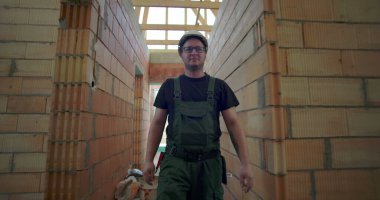 Yeşil tulumlu bir adam bitmemiş binanın koridorunda yürüyor, gözlüklü, odaklanmış bir ifade takınıyor, tuğla duvarları ve ahşap iskeleti var.