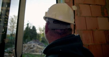 Sert şapkalı, açık havada duran, kapının yanında duran, arka planda doğal çevresi ve enkazı olan olgun inşaat işçilerinin yan profili.