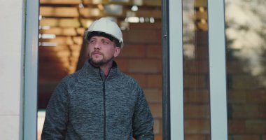 Kapıda duran inşaat işçisi, düşünceli bir şekilde mesafeye bakıyor, kafasında şapka, tuğla duvar arka planı, endüstriyi, işçiliği ve işçiliği temsil ediyor.