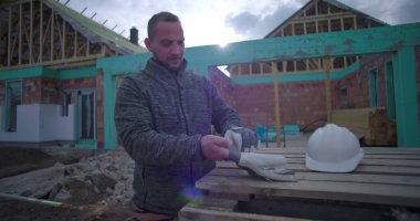 İnşaat alanında eldiven ve miğfer takan adam, arka planda kısmen inşa edilmiş evle ahşap paletlerin yanında duruyor, dışarıda çalışmaya hazırlanıyor.