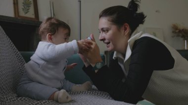 Anne bebekle kanepede iletişim kuruyor, şakacı yüz yüze iletişim kuruyor, erken çocukluk gelişiminde neşe ve bağlılık gösteriyor, ev ortamını büyütüyor.